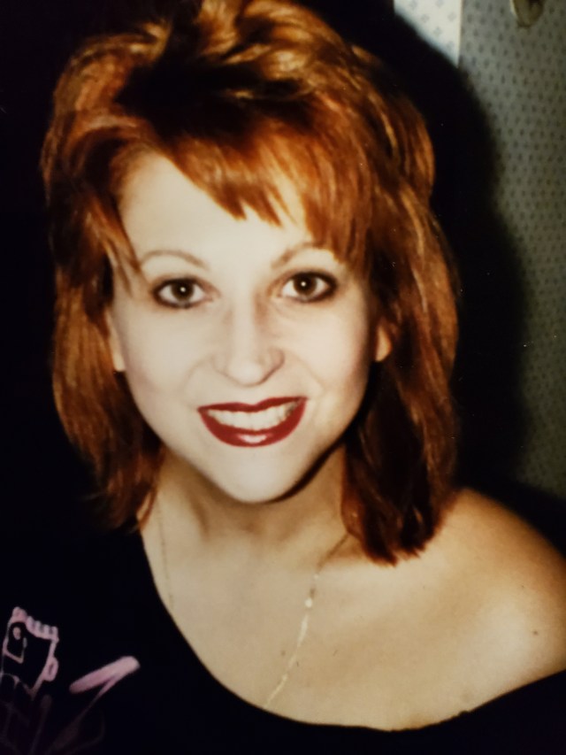 A portrait of a woman with vibrant red hair and a bright smile, wearing a black off-shoulder top, captured in a warm, nostalgic setting.