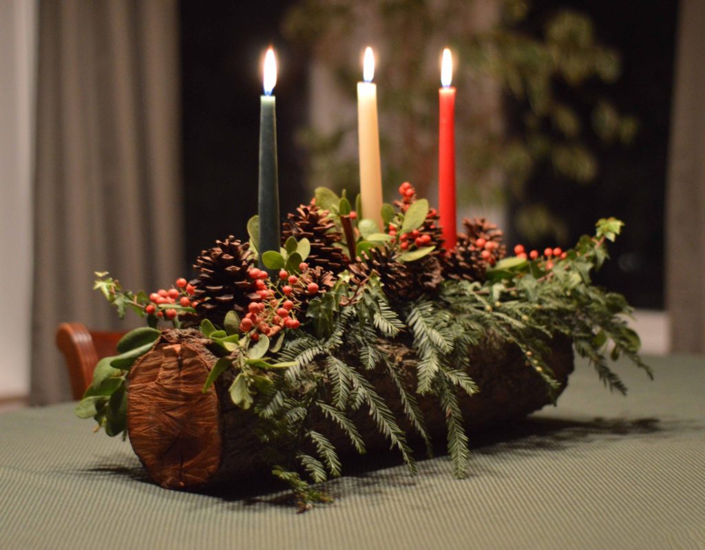 A decorated Yule log with three lit candles, pinecones, and greenery, placed on a table.