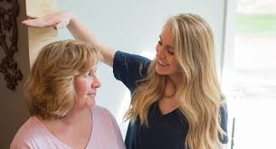 Two women in a bright room, one with longer blonde hair playfully measuring her height against the other woman with shorter, wavy hair.